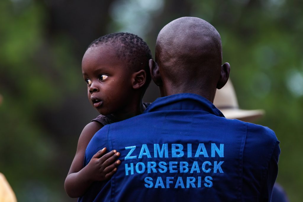 Picture of a man holding a child. The back of the mans shirt says Zambian Horseback Safaris 